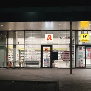 Eingangstür der Apotheke B29 am Abend mit Sicht in das Schaufenster und das Logo über der Tür